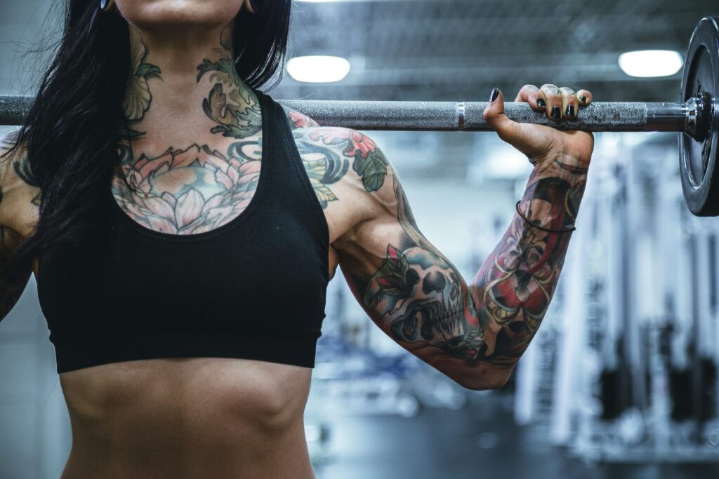 close up of tattooed woman in sports bra holding a barbell on her back.
