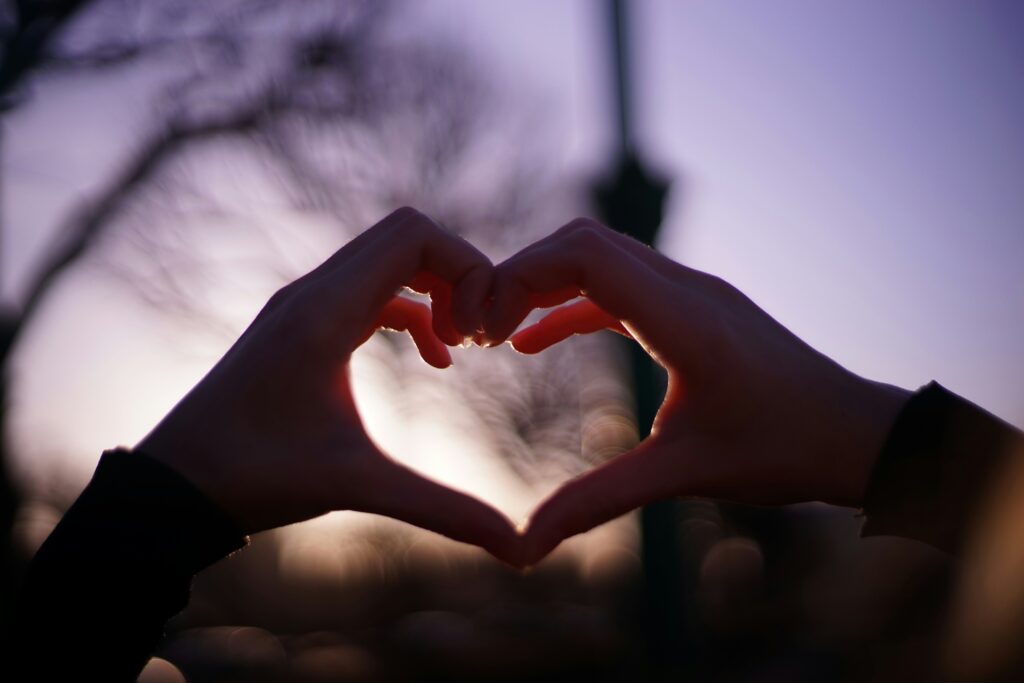 close up of hands making a heart shape in front of a sunset